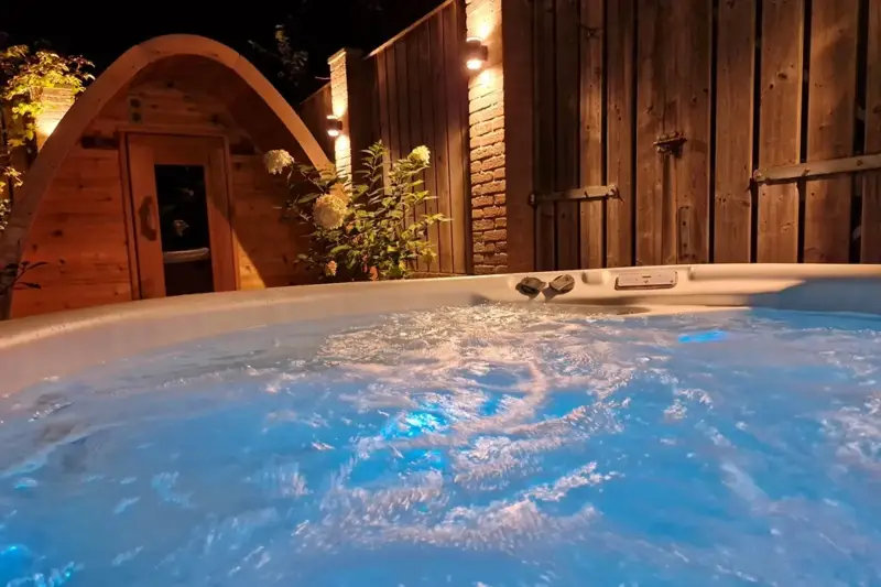 Bain nordique Nordic Hottub Classic bouillonnant et éclairé avec sauna Red Cedar Pod en arrière-plan le soir