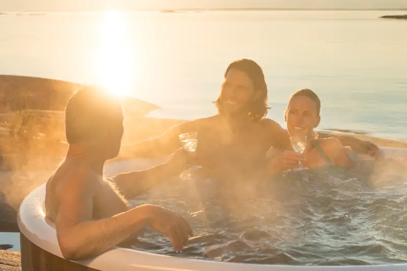 Des personnes profitant d'un verre dans le bain nordique Skargards Rojal avec poêle intégré au coucher du soleil