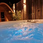 Bain nordique Nordic Hottub Classic bouillonnant et éclairé avec sauna Red Cedar Pod en arrière-plan le soir